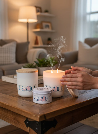 Three white candles with colorful numbers on the label are arranged on a wooden coffee table in a cozy living room. One candle is lit with visible flames and rising smoke, while a person's hands gently hold its handle.
