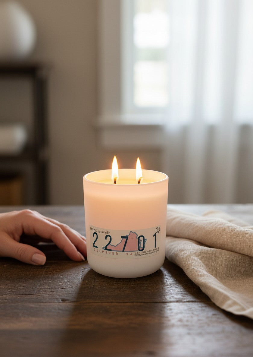 A lit white candle in a frosted glass jar sits on a wooden table, with a person's hand resting nearby and a beige cloth beside it. 