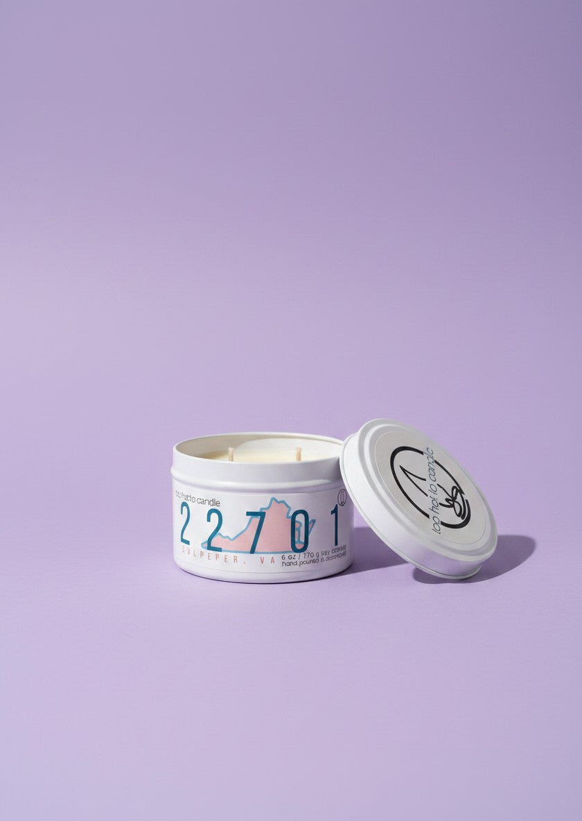 A white metal tin candle with its lid leaning against it on a solid lavender background. The candle features a colorful graphic of a mountain range with the numbers '2 2 7 0 1' printed on the front.